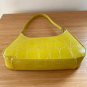Puede incluir: Un bolso amarillo brillante con textura de piel de cocodrilo. El bolso tiene un asa curva y forma rectangular. Est&aacute; hecho de un material brillante y tiene costuras blancas a lo largo de las costuras.