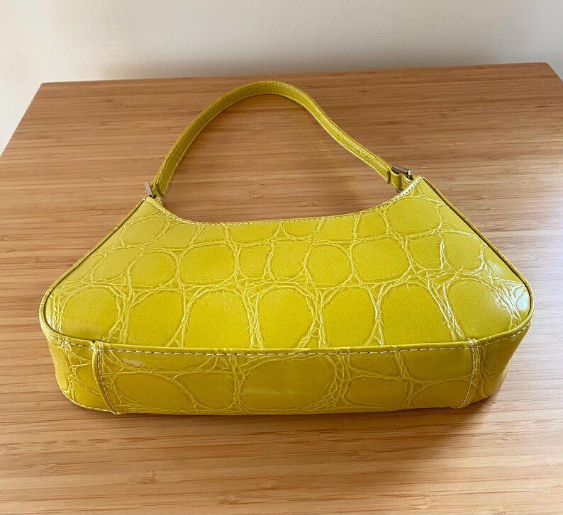 Puede incluir: Un bolso amarillo brillante con textura de piel de cocodrilo. El bolso tiene un asa curva y forma rectangular. Est&aacute; hecho de un material brillante y tiene costuras blancas a lo largo de las costuras.