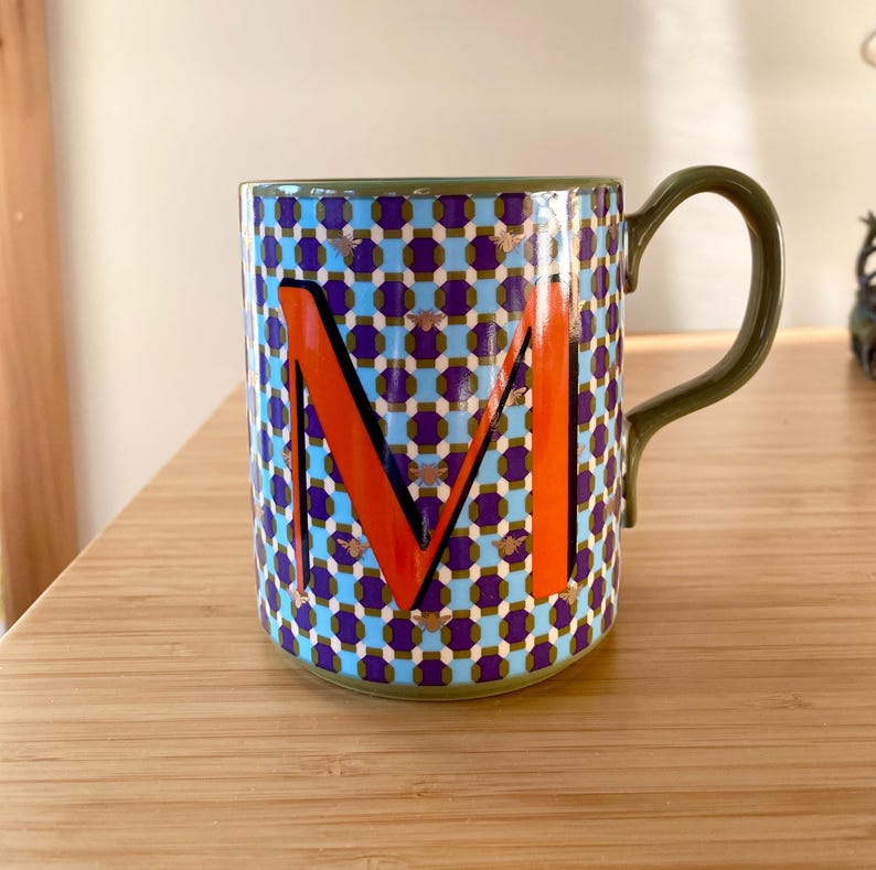 Puede incluir: Una taza de cer&aacute;mica con asa y borde verdes. La taza presenta un patr&oacute;n repetitivo de formas azules y moradas con detalles dorados. Una gran letra "M" naranja se muestra prominentemente en la parte delantera.
