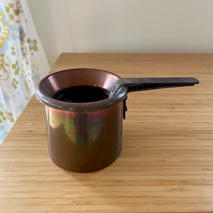 May include: A small copper-colored pot with a dark interior, likely containing a liquid. The pot has a short, flat handle and a rounded rim. The surface of the pot has a metallic sheen, reflecting various colors. The pot sits on a wooden surface.