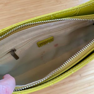 Puede incluir: Vista abierta de un bolso verde lima con un patr&oacute;n texturizado similar al de un cocodrilo. El interior es beige con una peque&ntilde;a etiqueta rectangular. Una cremallera plateada recorre la parte superior y una mano es visible en el lado izquierdo.