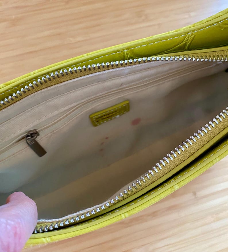 Puede incluir: Vista abierta de un bolso verde lima con un patr&oacute;n texturizado similar al de un cocodrilo. El interior es beige con una peque&ntilde;a etiqueta rectangular. Una cremallera plateada recorre la parte superior y una mano es visible en el lado izquierdo.