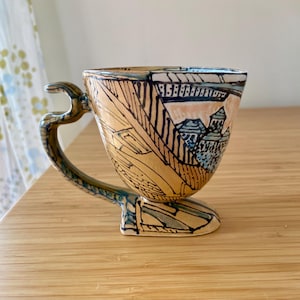 May include: A handcrafted ceramic mug with a unique, artistic design. The mug features a tan base color with black and blue line art depicting a landscape scene. The handle and base have a curved, organic shape. The mug is a decorative piece.