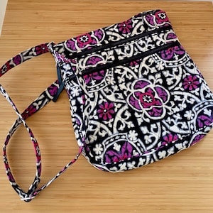 May include: A quilted crossbody bag featuring a black, white, and magenta floral-inspired geometric pattern. The bag includes an adjustable shoulder strap and two horizontal zippered compartments on the front, each with a black zipper pull.