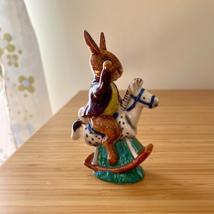May include: A ceramic figurine of a brown rabbit wearing a jacket, riding a white and black spotted rocking horse. The rocking horse has a green base and brown rockers. The figurine is on a wooden surface.