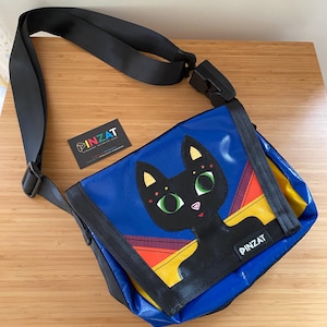 May include: A blue messenger bag featuring a black cat graphic with green eyes, yellow and red geometric accents, and a black adjustable shoulder strap. The bag has a black label with the white text PINZAT and includes a matching brand card.