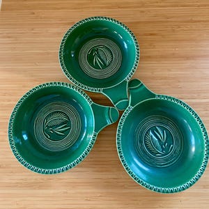May include: Three green ceramic bowls with handles. Each bowl features a white spiral design and a leaf motif in the center. The rims are decorated with a white zigzag pattern. The bowls are arranged on a wooden surface.