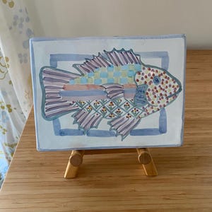 May include: A rectangular ceramic tile featuring a hand-painted fish design. The fish body is divided into sections with purple stripes, a blue and yellow checkered pattern, and a floral motif. The head is decorated with colorful dots.