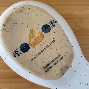 May include: White ceramic spoon rest with speckled design. Features a tan center with two yellow birds and blue floral accents. The text "by ANTHROPOLOGIE" is visible, along with "DISHWASHER & MICROWAVE SAFE" and "MADE IN CHINA".