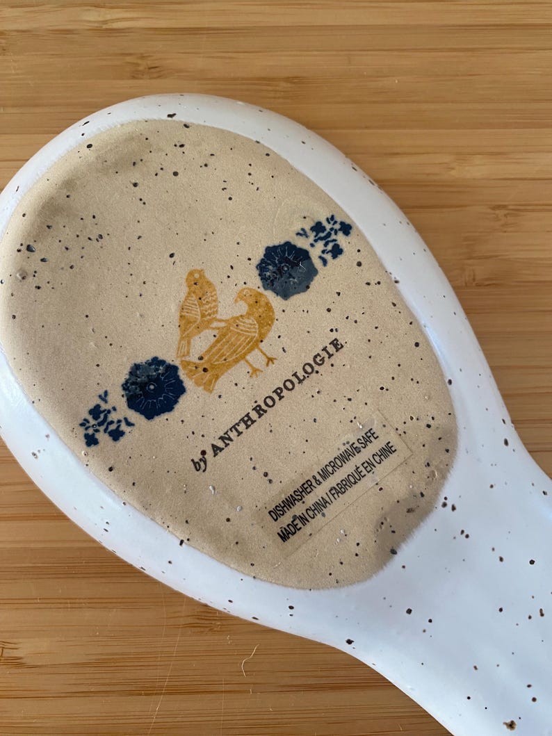 May include: White ceramic spoon rest with speckled design. Features a tan center with two yellow birds and blue floral accents. The text "by ANTHROPOLOGIE" is visible, along with "DISHWASHER & MICROWAVE SAFE" and "MADE IN CHINA".