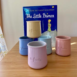 May include: Five ceramic pots in pastel shades of yellow, blue, light blue, and pink, each embossed with the text la fermière. The pots are arranged in front of a blue hardcover book titled The Little Prince by Antoine de Saint-Exupéry.