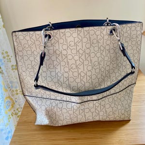 May include: A beige and white tote bag with a repeating "ck" pattern. The bag has a navy blue interior and handles, with silver-tone hardware. The bag is sitting on a wooden surface.