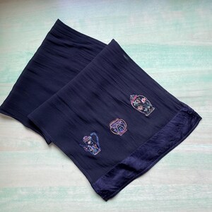 May include: Navy blue scarf with embroidered floral and teapot designs. The scarf has a textured appearance and a darker blue satin border. The embroidered designs are in shades of pink, blue, green, and purple.