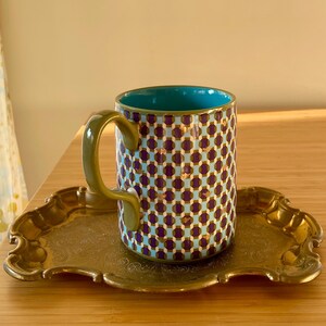 Puede incluir: Una taza de cer&aacute;mica estampada con interior turquesa y asa dorada se encuentra sobre una bandeja dorada. El exterior de la taza presenta un patr&oacute;n repetido de formas moradas y doradas sobre un fondo azul claro. La bandeja tiene un borde decorativo y adornado.