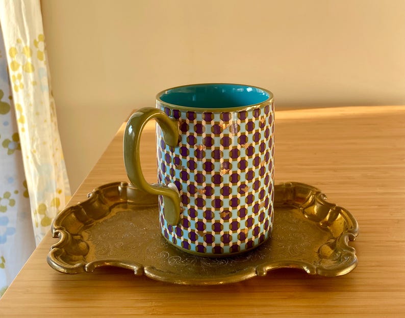 Puede incluir: Una taza de cer&aacute;mica estampada con interior turquesa y asa dorada se encuentra sobre una bandeja dorada. El exterior de la taza presenta un patr&oacute;n repetido de formas moradas y doradas sobre un fondo azul claro. La bandeja tiene un borde decorativo y adornado.
