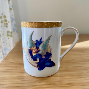 May include: White ceramic mug with a gold rim and handle. The mug features an illustration of an angel in blue robes and wings, surrounded by a golden halo. The design is centered on the mug's surface.