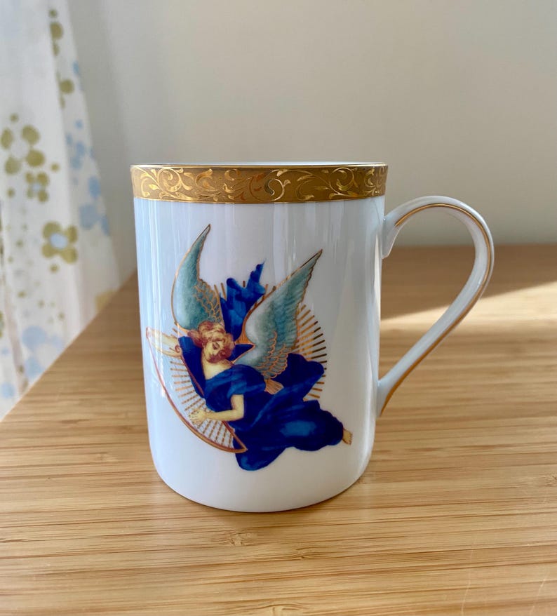 May include: White ceramic mug with a gold rim and handle. The mug features an illustration of an angel in blue robes and wings, surrounded by a golden halo. The design is centered on the mug's surface.
