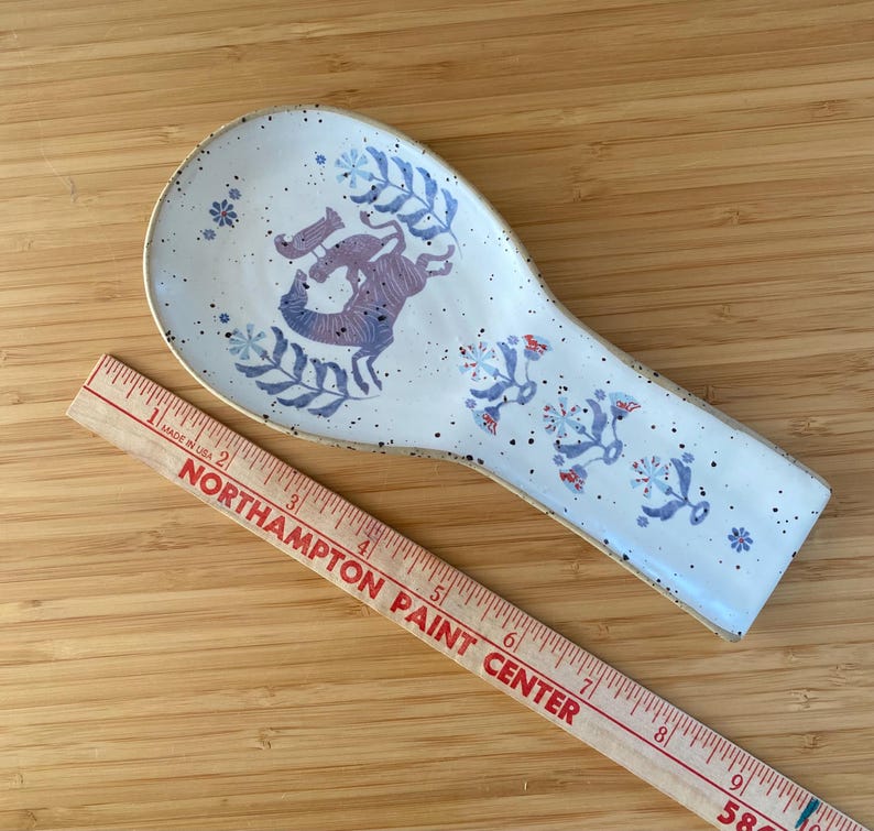 May include: A speckled white ceramic spoon rest with a light brown rim. The spoon rest features a whimsical design of purple animals and blue floral accents. A wooden ruler is in the foreground.