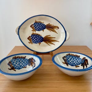 May include: A ceramic serving set consisting of one oval platter and two matching bowls. Each piece is off-white with a blue rim and features hand-painted fish motifs with blue scale patterns and brown fins, tails, and heads on the interior surfaces.