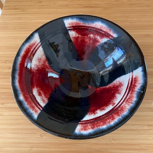 May include: A circular ceramic bowl featuring a glossy glaze with a radial pattern of deep black and vibrant red segments. The colors blend into white transitions at the edges, creating a high-contrast, multi-colored finish on the interior surface.