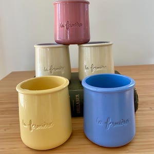 May include: Five colorful ceramic jars in shades of pink, yellow, cream, and blue are arranged on a wooden surface. Each jar has a cylindrical shape with a slightly flared base and the embossed text "la fermière".