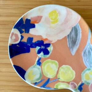 May include: A ceramic spoon rest with a floral design. The spoon rest features a peach-colored background with painted flowers in shades of white, yellow, blue, and pink. The spoon rest has a curved shape and a white rim.