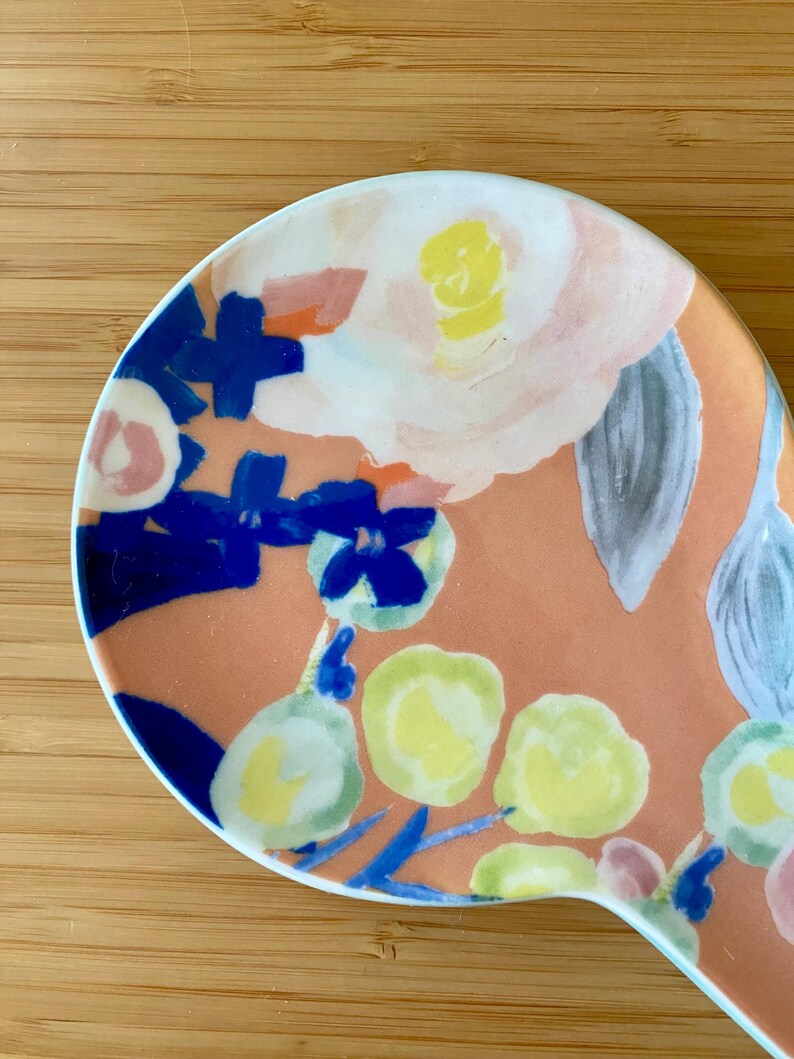 May include: A ceramic spoon rest with a floral design. The spoon rest features a peach-colored background with painted flowers in shades of white, yellow, blue, and pink. The spoon rest has a curved shape and a white rim.