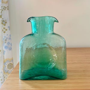 May include: A translucent teal glass vase with a square-shaped body and a wide, flared rim. The glass features a textured, slightly uneven surface with a prominent circular indentation centered on the front panel of the vessel.