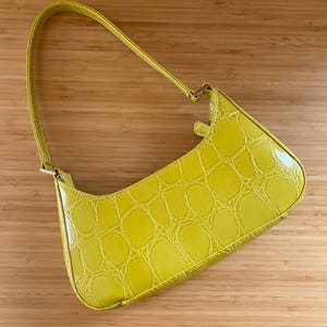 Puede incluir: Un bolso amarillo brillante con textura de piel de cocodrilo. El bolso tiene forma curva, un asa y un cierre dorado. El bolso est&aacute; hecho de un material brillante y se encuentra sobre un fondo de madera.