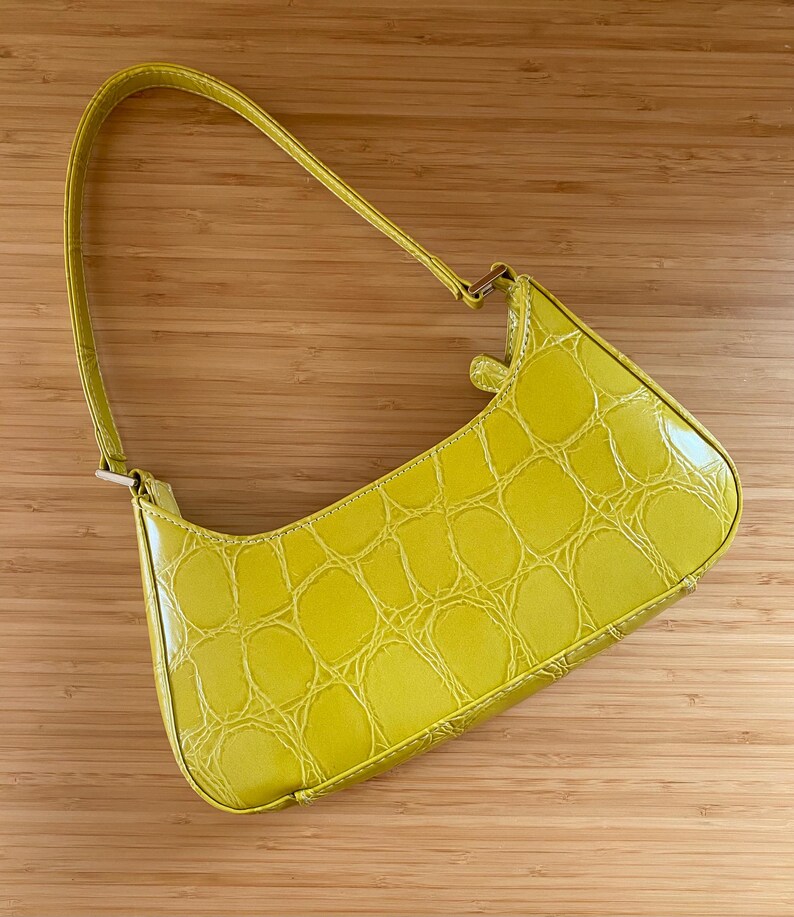 Puede incluir: Un bolso amarillo brillante con textura de piel de cocodrilo. El bolso tiene forma curva, un asa y un cierre dorado. El bolso est&aacute; hecho de un material brillante y se encuentra sobre un fondo de madera.