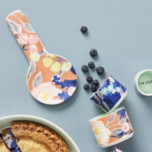 May include: Overhead shot of a ceramic spoon rest, measuring cups, and a pie. The spoon rest and measuring cups have a floral design with orange, blue, and yellow accents. A green measuring cup reads "1/4 CUP". Blueberries are scattered around the items.