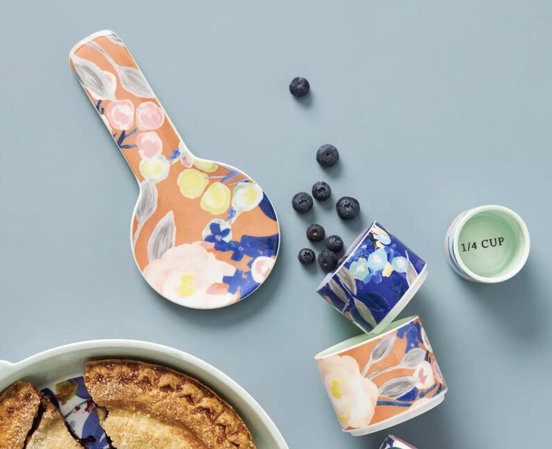 May include: Overhead shot of a ceramic spoon rest, measuring cups, and a pie. The spoon rest and measuring cups have a floral design with orange, blue, and yellow accents. A green measuring cup reads "1/4 CUP". Blueberries are scattered around the items.