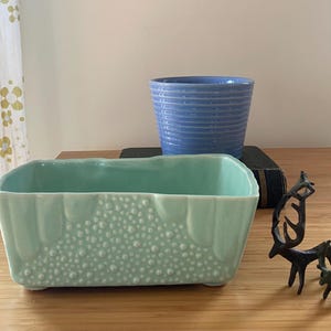 May include: A rectangular, light-green ceramic planter with a textured, dotted pattern and a light blue ceramic pot with horizontal ridges. Two small, dark metal deer figurines are also present.