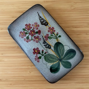 May include: Rectangular ceramic dish with a floral design. The dish has a gray background with a dark brown border. The floral design features red flowers, green leaves, and yellow accents. The dish is placed on a wooden surface.