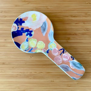 May include: A ceramic spoon rest with a peach background and a floral design in white, blue, yellow, and pink. The spoon rest has a rounded head and a long, handle-like extension.