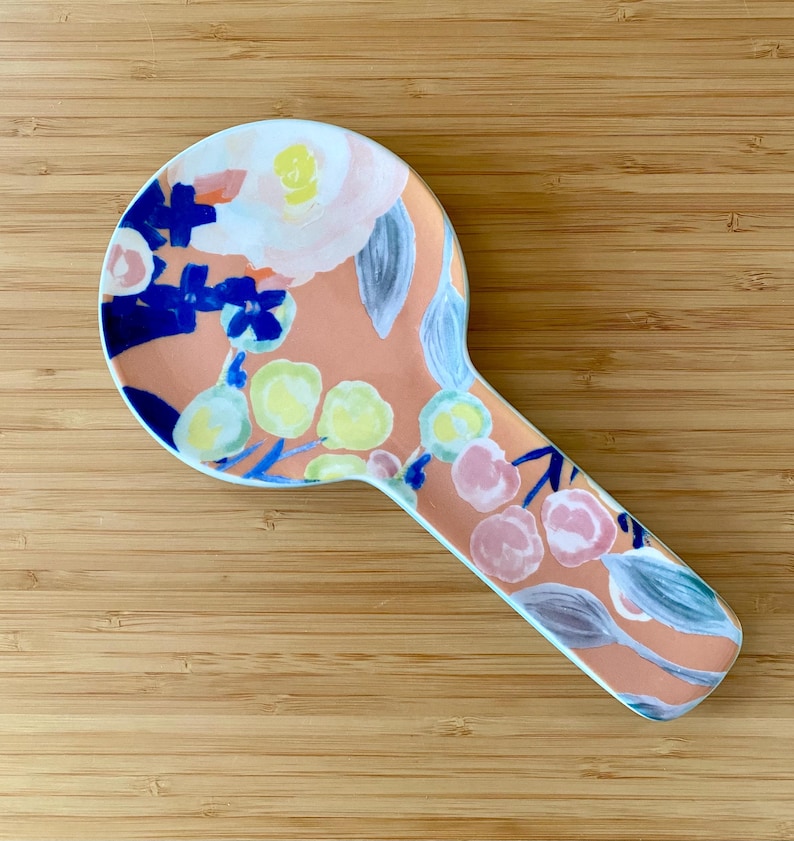 May include: A ceramic spoon rest with a peach background and a floral design in white, blue, yellow, and pink. The spoon rest has a rounded head and a long, handle-like extension.