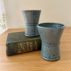 May include: Two handmade, speckled blue ceramic vases with a unique hourglass shape. The vases are displayed on a wooden surface next to a vintage book with gold lettering. The vases have a light blue rim and base.