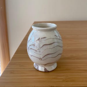 May include: A small, round ceramic vase with a glossy off-white glaze featuring horizontal, wavy brown marbled patterns. The vase has a narrow, circular opening at the top and a slightly flared base, showcasing a smooth, reflective surface texture.