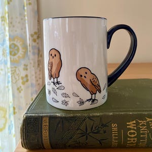 Może przedstawiać: Biały ceramiczny kubek z granatowym uchwytem i obramowaniem. Kubek przedstawia dwie brązowe ilustracje s&oacute;w z jesiennymi liśćmi. Kubek stoi na zielonej książce.