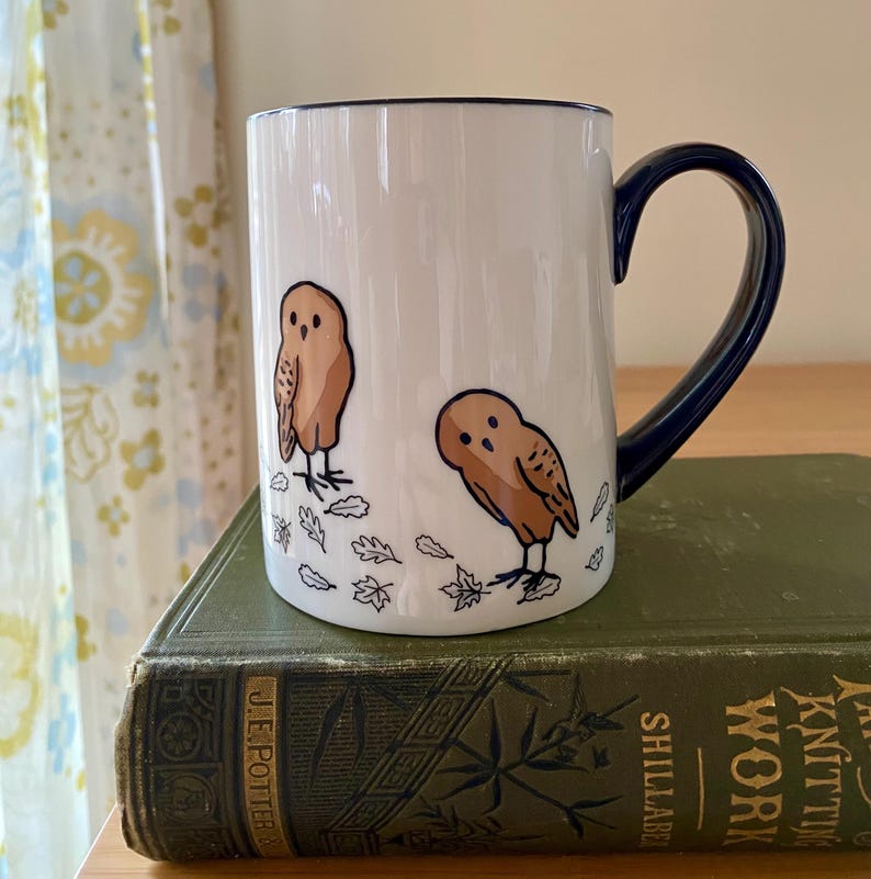 Może przedstawiać: Biały ceramiczny kubek z granatowym uchwytem i obramowaniem. Kubek przedstawia dwie brązowe ilustracje s&oacute;w z jesiennymi liśćmi. Kubek stoi na zielonej książce.