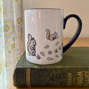 Może przedstawiać: Biały ceramiczny kubek z granatowym uchwytem i obramowaniem. Kubek przedstawia ilustracje dw&oacute;ch wiewi&oacute;rek i jesiennych liści. Kubek stoi na zielonej, zabytkowej książce.