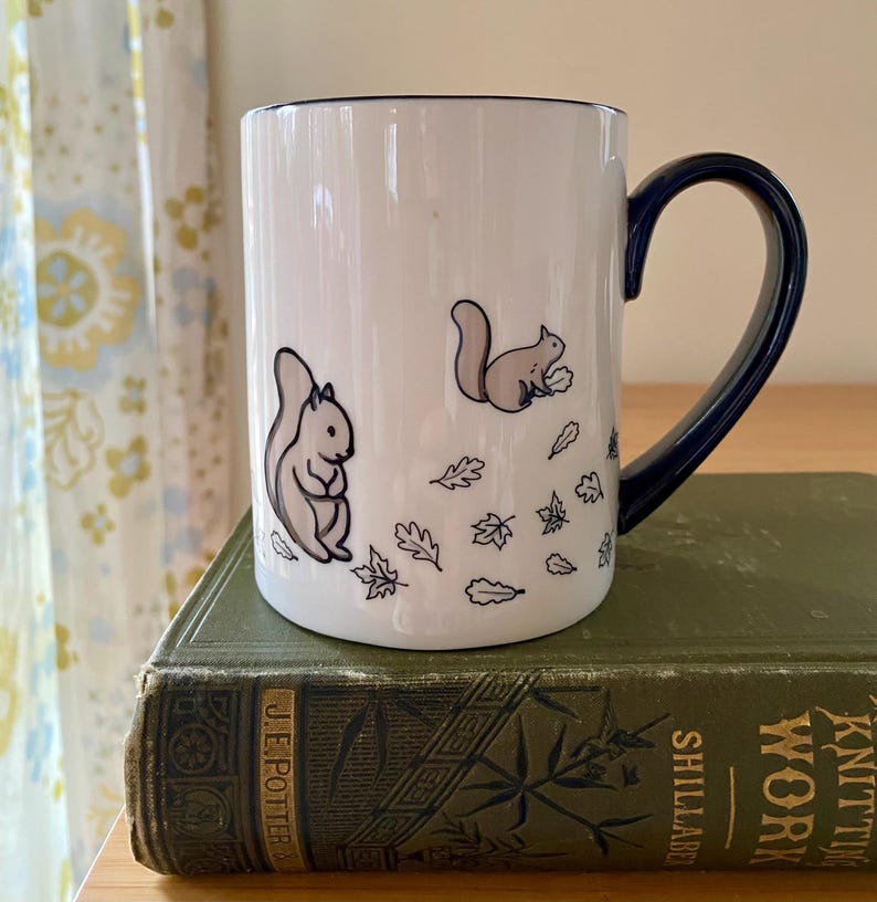 Może przedstawiać: Biały ceramiczny kubek z granatowym uchwytem i obramowaniem. Kubek przedstawia ilustracje dw&oacute;ch wiewi&oacute;rek i jesiennych liści. Kubek stoi na zielonej, zabytkowej książce.