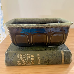 May include: A rectangular, brown ceramic dish with a green rim, resting on a vintage green book. The dish has a glossy finish and a series of curved indentations along its sides. The book's cover features gold lettering.