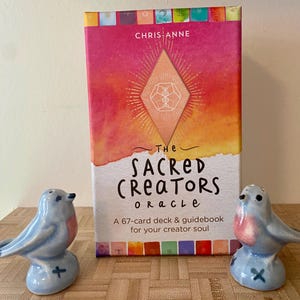 May include: A box for a 67-card deck of The Sacred Creators Oracle cards. The box is pink, orange, and yellow with a watercolor effect. The text on the box reads "The Sacred Creators Oracle" and "A 67-card deck & guidebook for your creator soul".