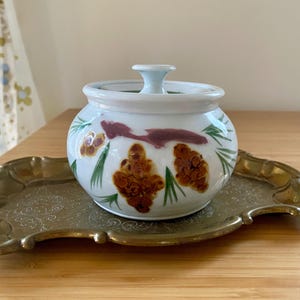 May include: A small, white ceramic jar with a lid, decorated with brown and yellow floral designs and green leaves. The jar sits on a decorative, gold-colored metal tray. The background is a light wood surface.