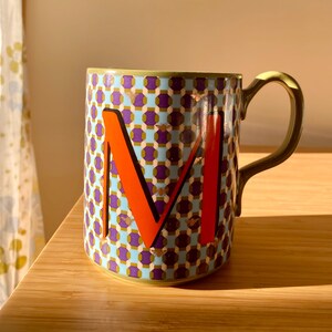 Puede incluir: Taza de cer&aacute;mica con un patr&oacute;n geom&eacute;trico en azul, morado y dorado. Una letra "M" naranja destaca en la parte delantera. El asa es verde oliva. La taza est&aacute; sobre una superficie de madera.