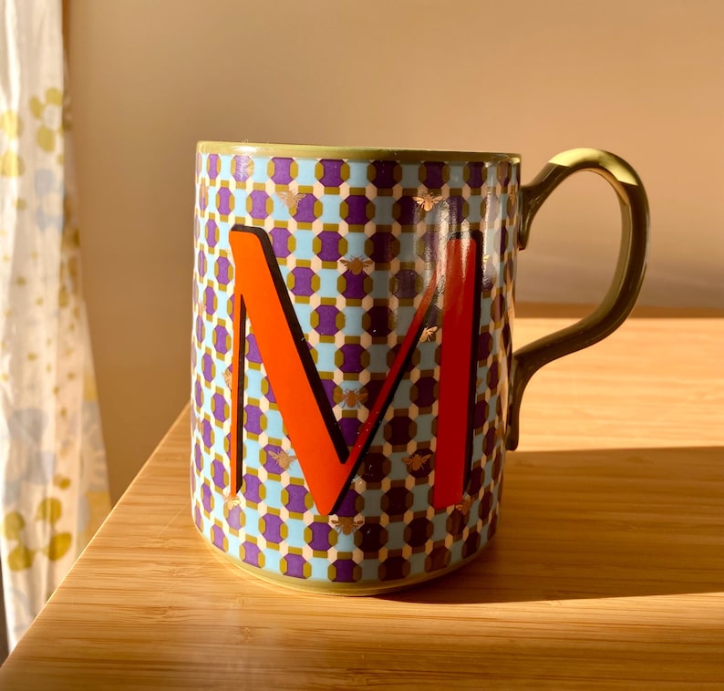 Puede incluir: Taza de cer&aacute;mica con un patr&oacute;n geom&eacute;trico en azul, morado y dorado. Una letra "M" naranja destaca en la parte delantera. El asa es verde oliva. La taza est&aacute; sobre una superficie de madera.