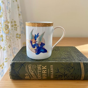 May include: White ceramic mug with a gold rim and handle, featuring a blue angel design. The mug is set on a vintage green book with gold lettering. The mug's design includes a winged figure in blue robes.