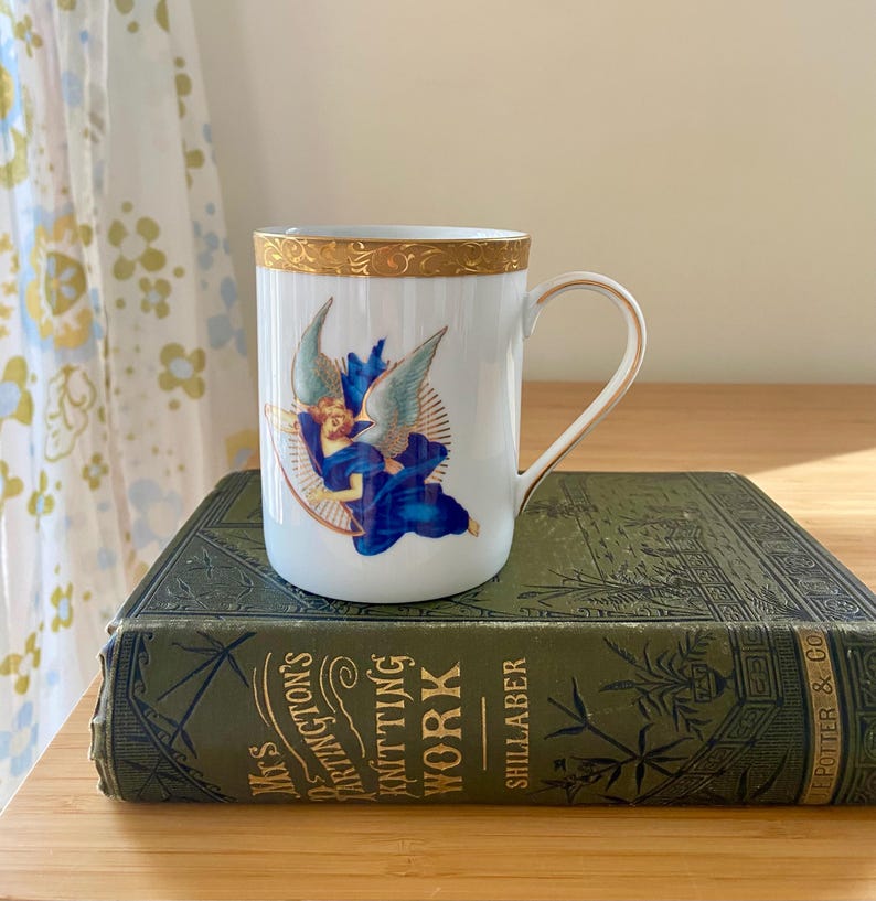 May include: White ceramic mug with a gold rim and handle, featuring a blue angel design. The mug is set on a vintage green book with gold lettering. The mug's design includes a winged figure in blue robes.