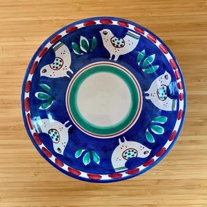 May include: A round, hand-painted ceramic bowl with a blue background. The bowl features white bird designs with green and red accents, and green leaf motifs. The center has a white interior with green and red concentric circles. The rim has a red and white pattern.